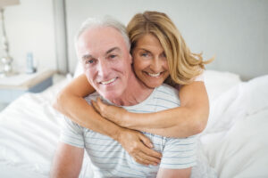 Smiling older couple sitting on a bed, with the woman hugging the man from behind.